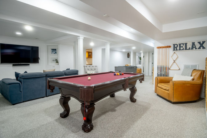 Finished home basement game room with burgundy-felt pool table, blue sectional sofa, leather armchair, wall cue rack and 'RELAX' sign, flat-screen TV and foosball table—ready for game night.