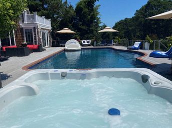 Sunny backyard oasis with bubbling white hot tub in the foreground, large blue swimming pool, wooden deck, lounge chairs, umbrellas and trees surrounding the space.