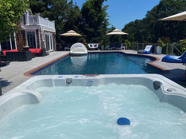 Sunny backyard oasis with bubbling white hot tub in the foreground, large blue swimming pool, wooden deck, lounge chairs, umbrellas and trees surrounding the space.