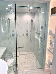 Bright spa-like bathroom with glass-enclosed walk-in shower, white marble walls, dual chrome rain showerheads and handheld wands, hexagon mosaic floor tile, built-in niche, and open shelving stocked with rolled towels.