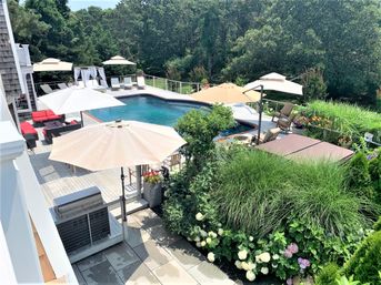 Sunny backyard pool with wooden deck, beige umbrellas, lounge chairs, red patio seating, hot tub cover and grill, surrounded by lush landscaping with hydrangeas, ornamental grasses and wooded backdrop