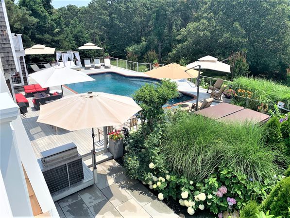 Sunny backyard pool with wooden deck, beige umbrellas, lounge chairs, red patio seating, hot tub cover and grill, surrounded by lush landscaping with hydrangeas, ornamental grasses and wooded backdrop