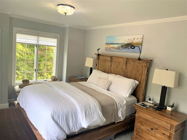 Bright master bedroom with king bed, crisp white linens and beige throw, rustic wooden headboard, matching nightstands, coastal canvas art and a window view of green trees.