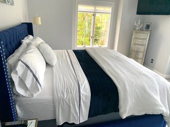 Bright modern bedroom with navy tufted upholstered bed, crisp white sheets with navy trim, navy velvet bed runner and textured white throw, sunlit window with green tree view and a silver tall dresser.