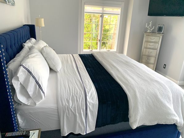 Bright modern bedroom with navy tufted upholstered bed, crisp white sheets with navy trim, navy velvet bed runner and textured white throw, sunlit window with green tree view and a silver tall dresser.