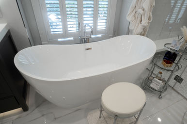 Spa-like modern bathroom with glossy white freestanding bathtub, chrome floor-mounted faucet, round cushioned stool, glass rolling cart stocked with toiletries, plush robe, and white plantation shutters over marble tile floor.
