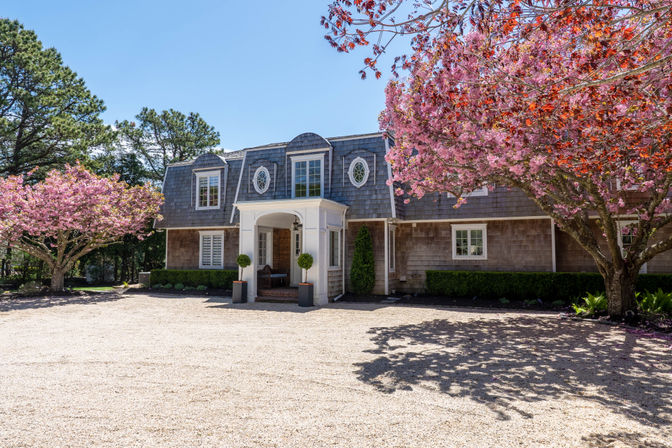 Sunny spring scene of a coastal shingle-style home with gambrel roof and white portico, gravel driveway framed by pink cherry blossom trees