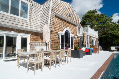 Coastal shingle-style house with a bright white poolside deck, weathered wooden dining table and chairs, umbrellas, wicker lounge seating with red cushions, sliding glass doors and arched entry, blue pool water and trees under a sunny sky.