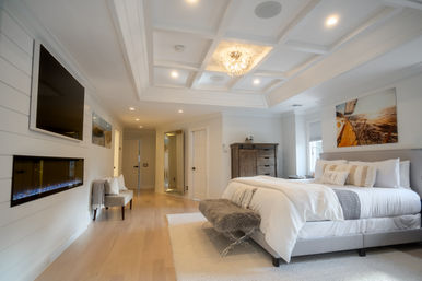 Bright coastal-modern master bedroom with tray ceiling and chandelier, king bed with white linens and fur bench, wooden dresser, wall-mounted TV above linear electric fireplace, light hardwood floors and sailboat wall art