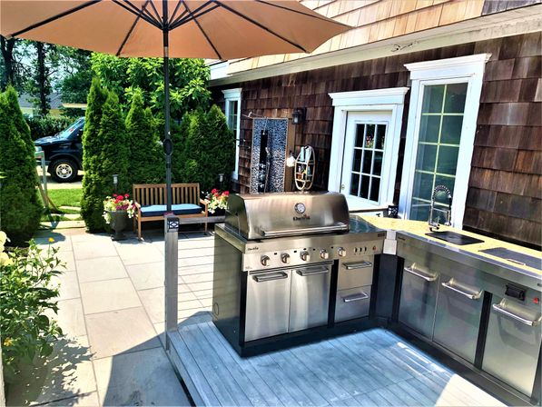 Sunny suburban backyard patio with stainless-steel outdoor kitchen and gas grill, built-in sink and stone countertops under a large umbrella, wooden bench with blue cushions, potted flowers and tall evergreen privacy hedges by a cedar-shingled house.