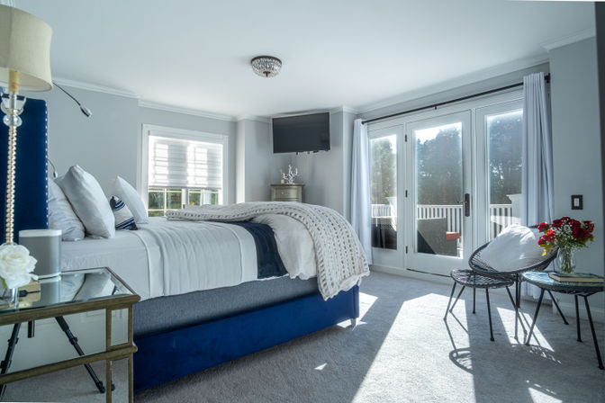 Sunlit modern master bedroom with blue upholstered bed, chunky knit throw, wall TV, glass French doors to a balcony and a small seating area with red roses.