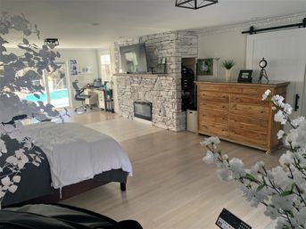 Spacious bright bedroom with light hardwood floors, white bedding on a platform bed, stacked stone fireplace with wall-mounted TV, large wooden dresser and small home office by sliding glass doors overlooking a backyard pool, decorative white blossoms in the foreground.