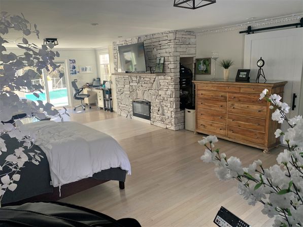 Spacious bright bedroom with light hardwood floors, white bedding on a platform bed, stacked stone fireplace with wall-mounted TV, large wooden dresser and small home office by sliding glass doors overlooking a backyard pool, decorative white blossoms in the foreground.