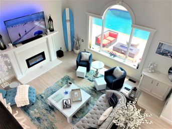 Bird's-eye view of a modern coastal living room with tufted sofa, blue accent chairs, marble fireplace, surfboard by a large arched window overlooking a bright blue pool and red patio seating