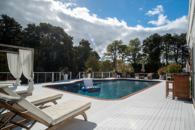 Inviting backyard swimming pool on a white painted wood deck with cushioned lounge chairs, draped canopy, blue inflatable float, and tree-lined yard under a partly cloudy sky.
