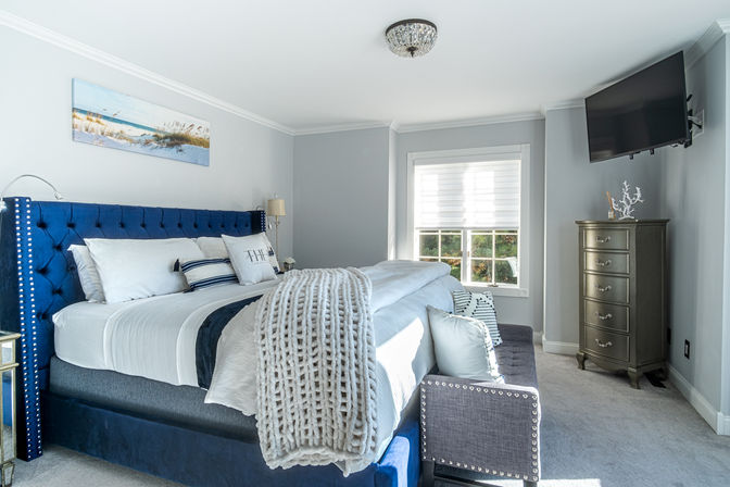 Sunlit coastal-inspired master bedroom with navy tufted king bed, chunky knit throw and striped pillows, gray walls, large window, wall-mounted TV and tall dresser
