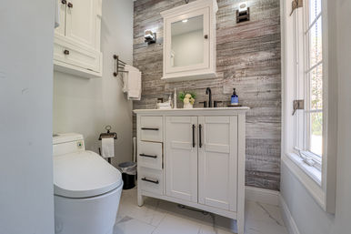 Cozy bright bathroom with white shaker vanity and mirrored medicine cabinet, reclaimed wood plank accent wall, matte black faucet and fixtures, modern toilet, towel rack and window with natural light.
