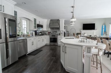 Bright modern white kitchen with large quartz island, stainless steel appliances, pendant lights, dark hardwood floors and a surfboard accent in an open-concept dining area.