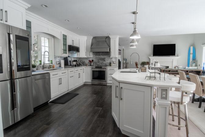Bright modern white kitchen with large quartz island, stainless steel appliances, pendant lights, dark hardwood floors and a surfboard accent in an open-concept dining area.