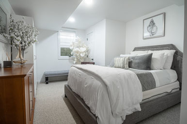 Bright modern bedroom with a gray upholstered bed, white duvet and pillows, wooden dresser with sailboat decor, tufted bench and large floral accents