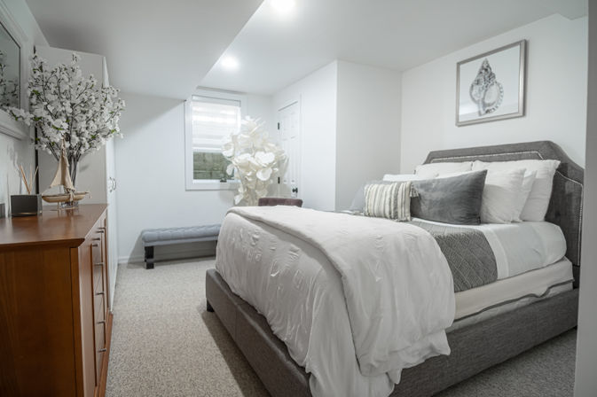 Bright modern bedroom with a gray upholstered bed, white duvet and pillows, wooden dresser with sailboat decor, tufted bench and large floral accents