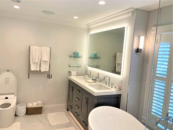 Bright modern master bathroom with gray double-sink vanity, backlit mirror, chrome faucets, freestanding white tub, wall-mounted towel rack with white towels, glass shelves, toilet and plantation-shutter window.