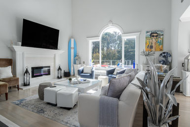 Sunlit modern coastal living room with large arched window and pool view, white marble fireplace with mounted TV, blue accent chairs, surfboard and glass coffee table.