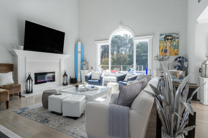 Sunlit modern coastal living room with large arched window and pool view, white marble fireplace with mounted TV, blue accent chairs, surfboard and glass coffee table.