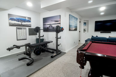 Cozy finished basement rec room with home gym — weight bench, barbell and dumbbells on rubber mats, exercise poster and canvas wall art; adjacent game area with red-felt pool table, blue sectional couch and wall-mounted TV.
