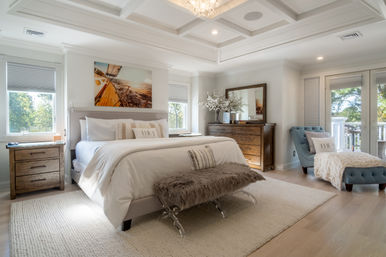 Sunlit coastal-style master bedroom with king bed and white linens, faux-fur bench, wooden nightstands and dresser, blue tufted chaise by balcony with tree views