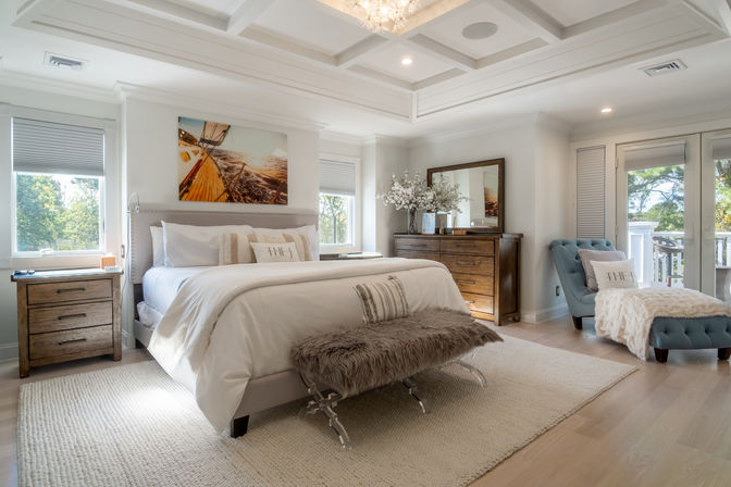 Sunlit coastal-style master bedroom with king bed and white linens, faux-fur bench, wooden nightstands and dresser, blue tufted chaise by balcony with tree views