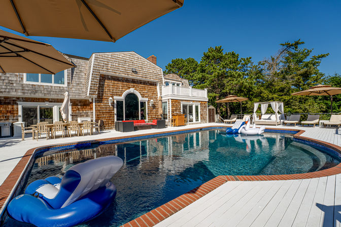 Coastal shingle-style beach house with brick-edged swimming pool, inflatable swan and blue float, white deck, umbrellas, cabana and lounge chairs under a clear blue sky.