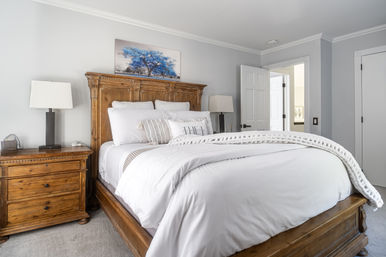 Bright master bedroom with a carved wooden bed frame, crisp white bedding and chunky knit throw, matching wooden nightstand with lamp, gray walls and blue tree artwork