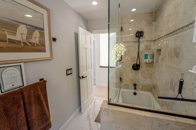 Bright modern bathroom with beige stone-tiled shower and bathtub behind a glass door, dark bronze fixtures, wall-mounted soap dispensers, brown towels, beach-chair artwork, and a white door opening to the hallway.