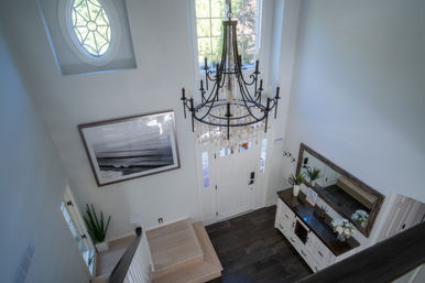 Sunlit two-story foyer from above with a wrought-iron crystal chandelier over a white paneled front door with sidelights, dark wood console and mirror, coastal black-and-white wall art, round decorative window and light wood staircase.