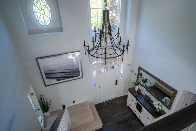 Sunlit two-story foyer from above with a wrought-iron crystal chandelier over a white paneled front door with sidelights, dark wood console and mirror, coastal black-and-white wall art, round decorative window and light wood staircase.