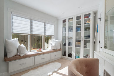 Sunlit modern dressing room with cushioned window seat and white pillows, built-in drawers, glass-front cabinets holding shoes and accessories, blush velvet chair and white faux-fur rug — airy neutral storage and seating area.