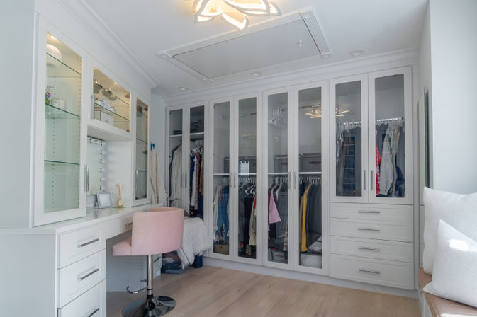 Bright modern walk-in closet and dressing room with glass-front built-in wardrobes, organized hanging clothes, white vanity with lighted mirror and a pink velvet swivel chair on light hardwood floors.