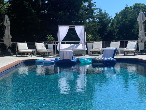 Sunny backyard pool deck with sparkling turquoise water, white canopy daybed and cushioned lounge chairs, blue inflatable floats drifting in the pool, and leafy trees in the background.