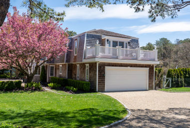 Shingle-style coastal house with white second-floor balcony and two-car garage, front yard featuring a blooming pink cherry tree, green lawn and gravel driveway on a sunny spring day.