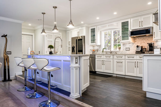 Bright modern white kitchen with island and blue LED-lit base, three white swivel bar stools, stainless steel appliances, pendant lights, subway tile backsplash and dark hardwood floors