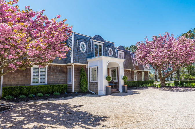 Shingle-style house with white columned portico, flanked by vibrant pink cherry blossom trees and trimmed hedges, gravel driveway under a bright blue spring sky.