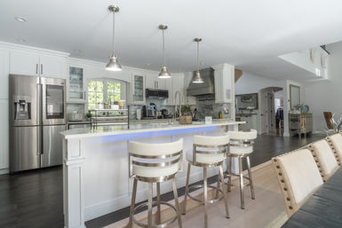 Bright modern open-concept kitchen with white cabinetry, large island with blue LED accent, three stainless-steel bar stools, pendant lights, stainless-steel French-door refrigerator and gray subway tile backsplash.