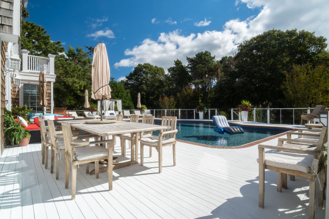 Sunny coastal-style backyard pool deck with white painted wood flooring, weathered wooden dining set, beige umbrellas, lounge chairs and a blue inflatable float
