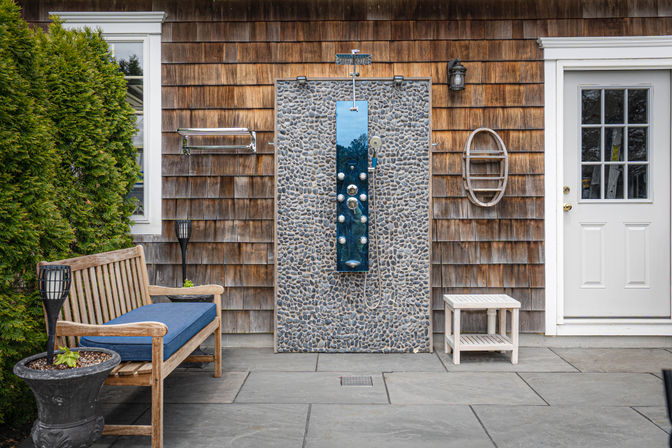 Outdoor pebble-tile shower panel with chrome fixtures mounted on cedar-shingle exterior, flanked by a white door, window and wooden bench with blue cushion on a stone patio.