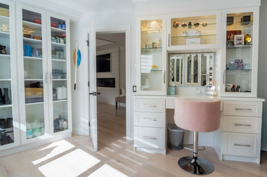 Bright modern dressing room with white built-in cabinets and glass-front shelving, light wood floor, illuminated makeup vanity with bulb mirror and pink velvet swivel stool bathed in natural light.