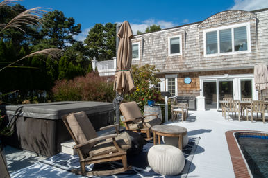 Sunlit backyard deck at a coastal-style shingle house featuring a covered hot tub, cushioned rocking chairs and small wooden table, knitted pouf, closed umbrella, outdoor dining set, grill and a pool under a blue sky.
