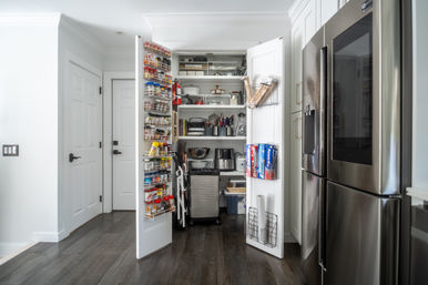 Open white kitchen pantry with organized shelves, door spice racks, and stainless-steel refrigerator on dark hardwood floors