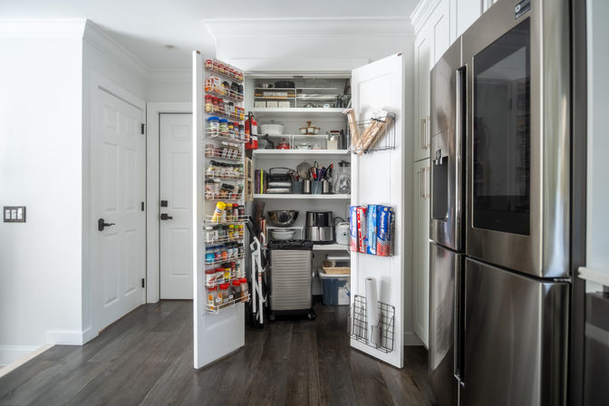 Open white kitchen pantry with organized shelves, door spice racks, and stainless-steel refrigerator on dark hardwood floors