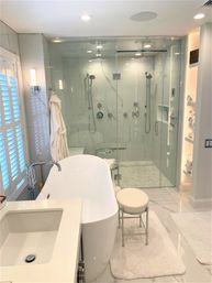 Spa-like modern master bathroom featuring a white freestanding tub, glass-enclosed dual rain shower with marble-look tile, vanity sink, plush rugs and stacked towels.